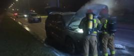 Strażacy gaszą samochód osobowy. Pożar miał miejsce pod maską pojazdu. Strażacy mają założone aparaty OUO i rozwiniętą linię wężową. W tle widać inne samochody.