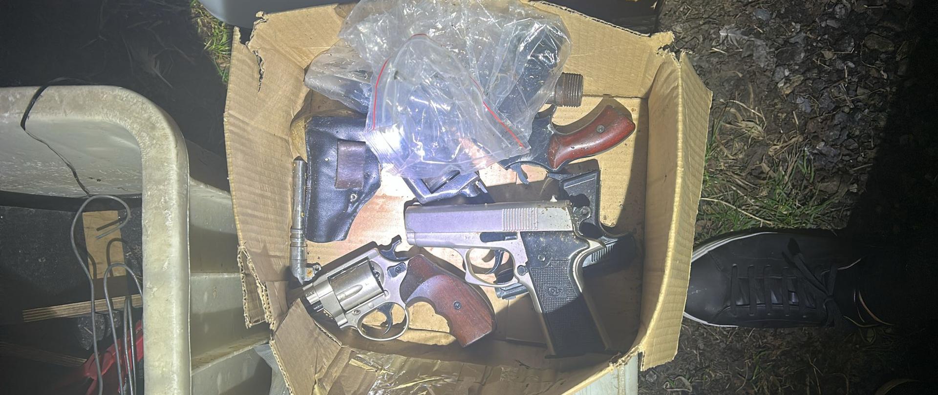 Otwarty karton, w środku widoczne pistolety i drobne akcesoria ułożone luzem.