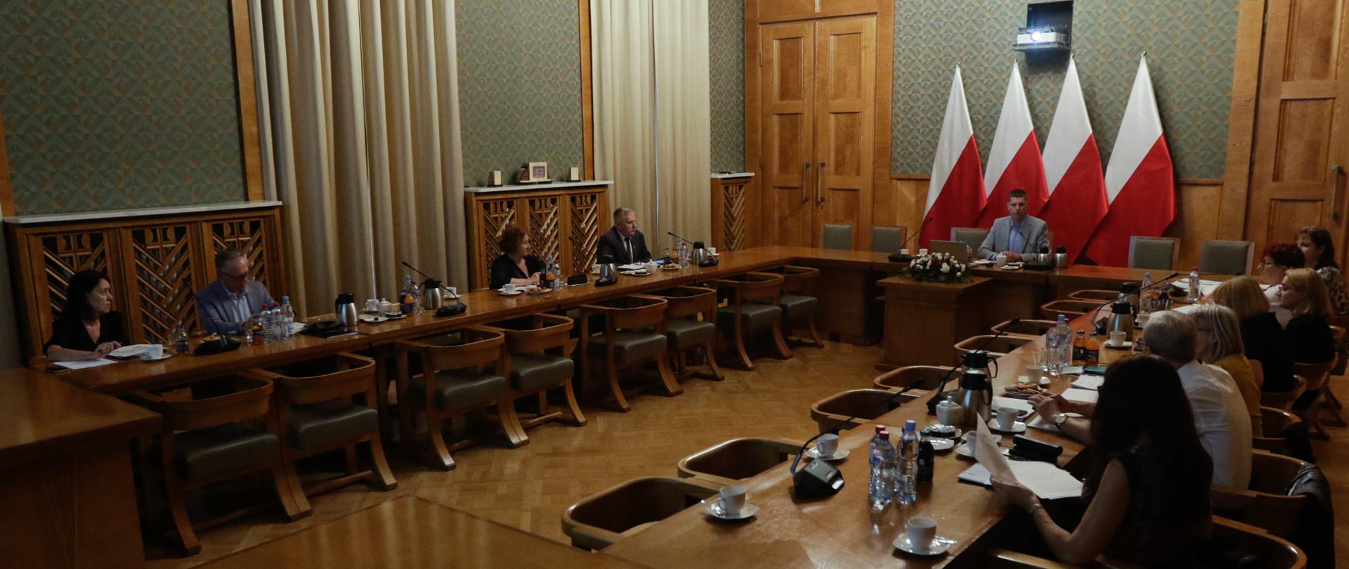 Ludzie siedzący przy stołach w dużej sali. W centrum wiceminister edukacji i nauki Dariusz Piontkowski na tle flag Polski