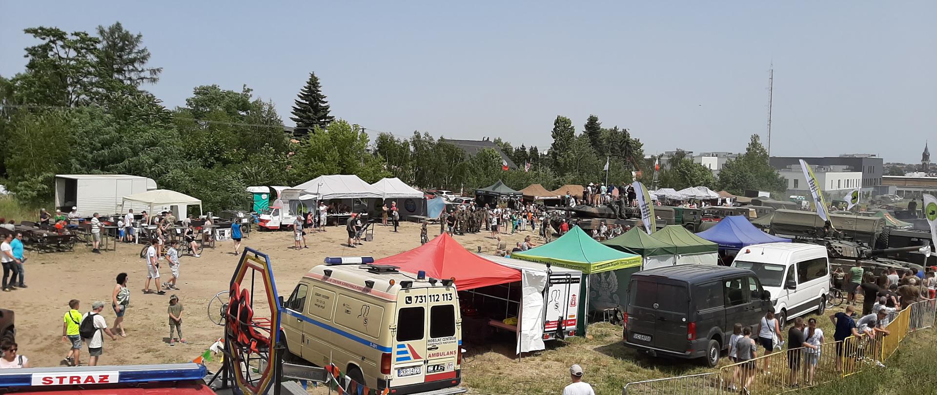 Na zdjęciu widać plac z dużą ilością pojazdów cywilnych oraz militarnych. Namioty wystawowe oraz osoby cywilne. Niebo błękitne. Słonecznie. W tle tereny zielone, zarośla.