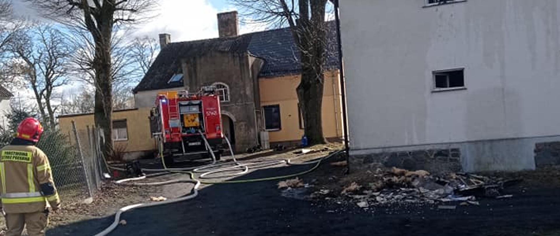 Zdjęcie przedstawia strażaka oraz samochód gaśniczy przed obiektem objętym pożarem