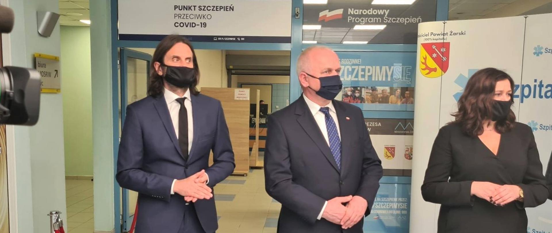 Wojewoda, dyrektor NFZ i prezes szpitala stoją w miejscu wygrodzonym. Za nimi ścianka promocyjna oraz wejście na teren pływalni