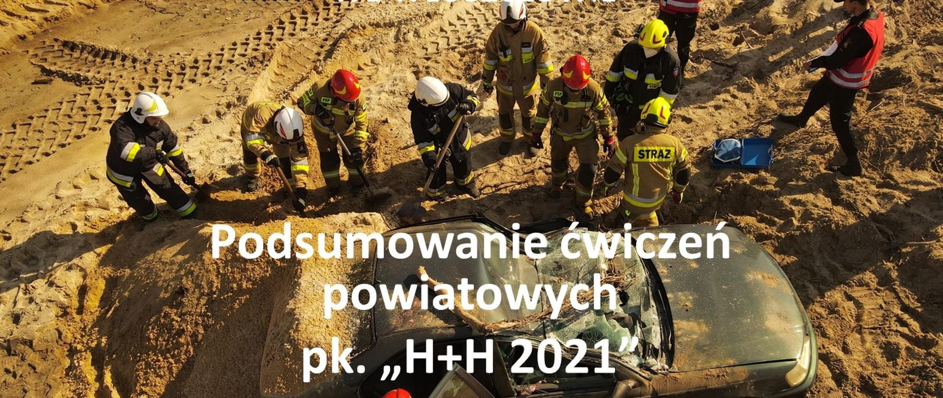 Kadr z ćwiczeń zrealizowany z drona, epizod II, samochód po dachowaniu z uszkodzonym dachem, częściowo przysypany piaskiem, wokół samochodu strażacy wykonujący dostęp do osoby w pojeździe, w górnej części fotografii napis Komenda Powiatowa PSP we Włoszczowie, poniżej tytuł podsumowanie ćwiczeń powiatowych H+H 2021
