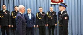 Delegacja strażaków PSP w umundurowaniu galowym podczas uroczystości wręczenia odznaczeń; na pierwszym planie strażak w galowym mundurze stoi naprzeciw Podsekretarza Stanu MSWiA Wiesława Leśniakiewicza, w tle poczet sztandarowy i funkcjonariusze PSP.
