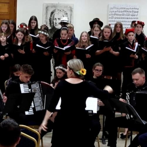 Na zdjęciu widać występ muzyczny w sali koncertowej. Na pierwszym planie dyrygentka prowadzi orkiestrę złożoną głównie z akordeonistów i skrzypków. W tle stoi duży chór dzieci i młodzieży ubrany w ciemne stroje, część osób ma na głowach tradycyjne wianki. W sali ustawiono krzesła dla publiczności, a za wykonawcami znajdują się obrazy i fortepian. Atmosfera wydarzenia jest uroczysta i artystyczna.