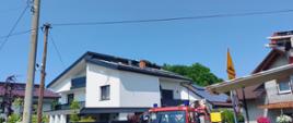 Widok ogólny. Widoczny dach budynku mieszkalnego jednorodzinnego po pożarze. Na dachu znajduje się instalacja fotowoltaiczna oraz panele solarne. Na bliższym planie samochody pożarnicze.