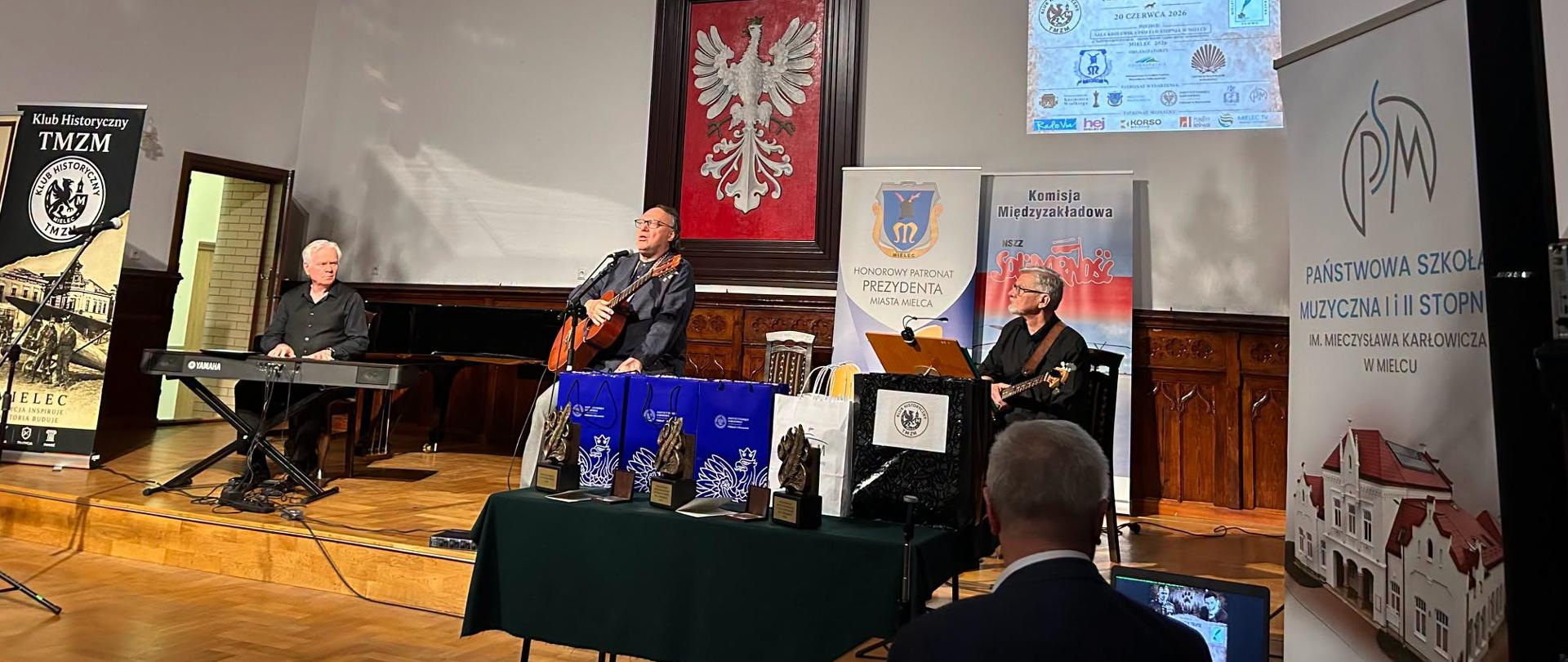 Trzech muzyków — bard z gitarą, klawiszowiec i gitarzysta basowy — występuje na scenie Sali Królewskiej podczas festiwalu „Na Wilczym Tropie", w tle herb Polski, freski z królami i banery organizatorów.
