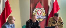 Jeden mężczyzna cywilny od lewo i dwóch funkcjonariuszy PSP siedzą przy stole prowadząc wideokonferencję