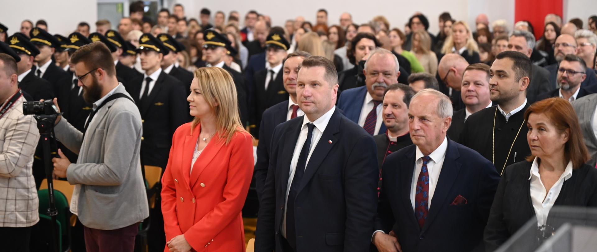 Widok od przodu na wypełnioną stojącymi ludźmi salę, w pierwszym rzędzie minister Czarnek i kobieta w pomarańczowym garniturze.