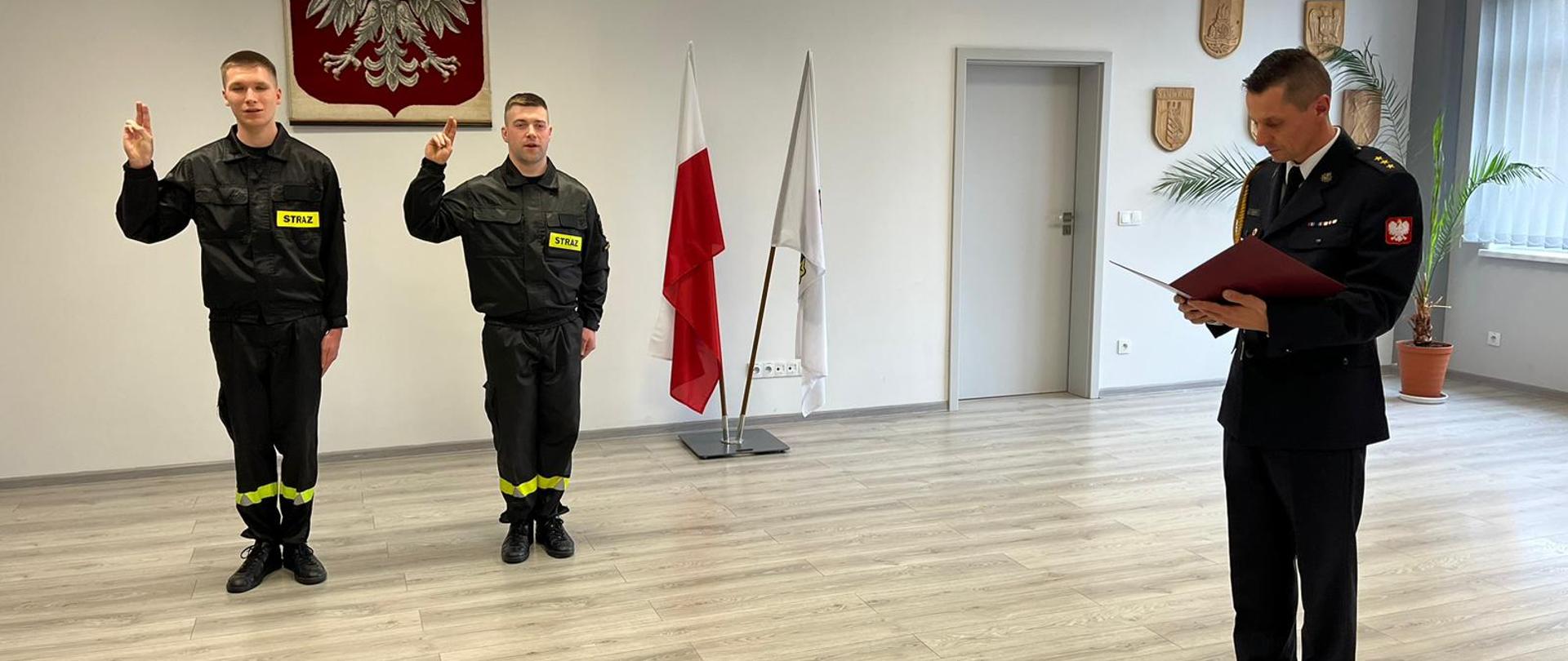 Na zdjęciu na środku nowo przyjęci funkcjonariusze składający ślubowanie, w tle Godło oraz flaga państwowa oraz flaga Państwowej Straży Pożarnej. Po prawej stronie stoi przyjmujący ślubowanie Komendant. 