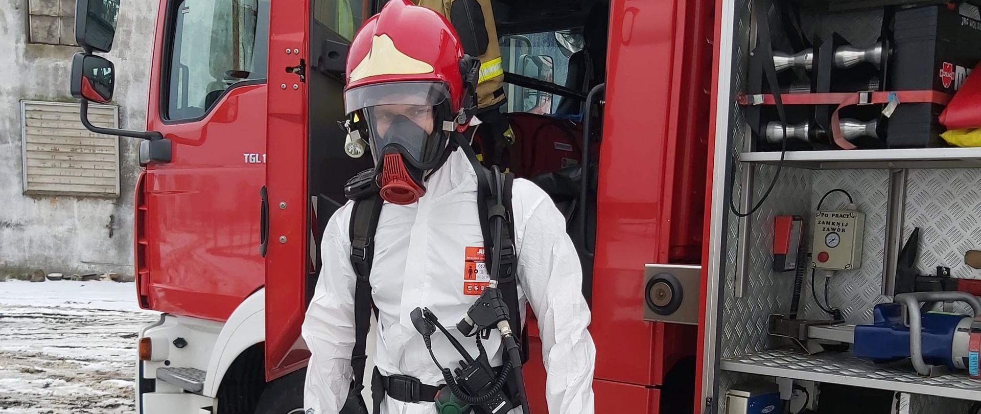 Strażak na tle wozu strażackiego. Ubrany w kombinezon, aparat powietrzny, kalosze.