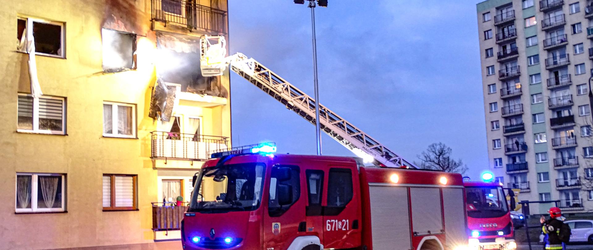 Na zdjęciu widoczny jest budynek wielorodzinny na osiedlu, mieszkanie na drugim piętrze z widocznymi uszkodzeniami po wybuchu i pożarze. Uszkodzona elewacja budynku. Przed budynkiem stoi samochód straży pożarnej średni ratowniczo-gaśniczy i samochód specjalny drabina mechaniczna z rozłożoną drabiną sięgającą koszem do drugiego piętra. 