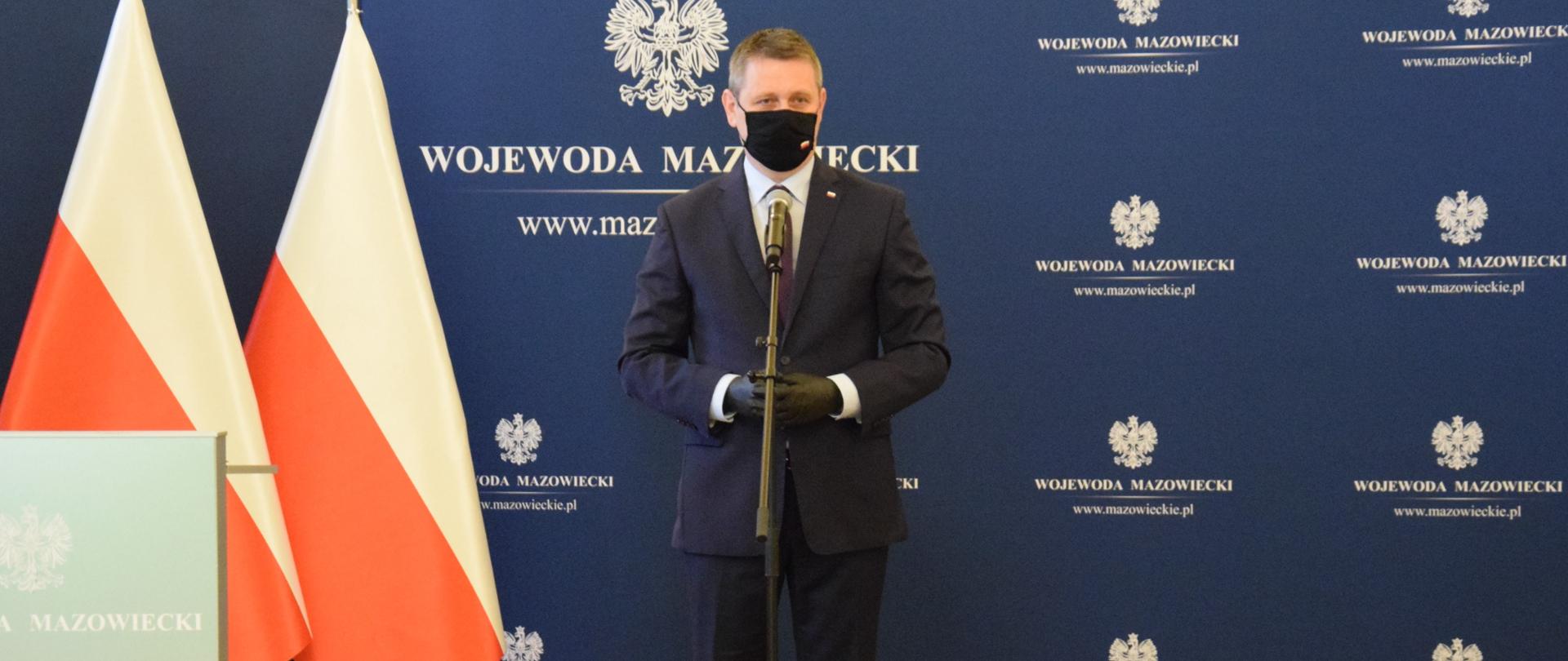 Na zdjęciu znajduje się mężczyzna, który mówi do mikrofonu, w tle flagi Polski