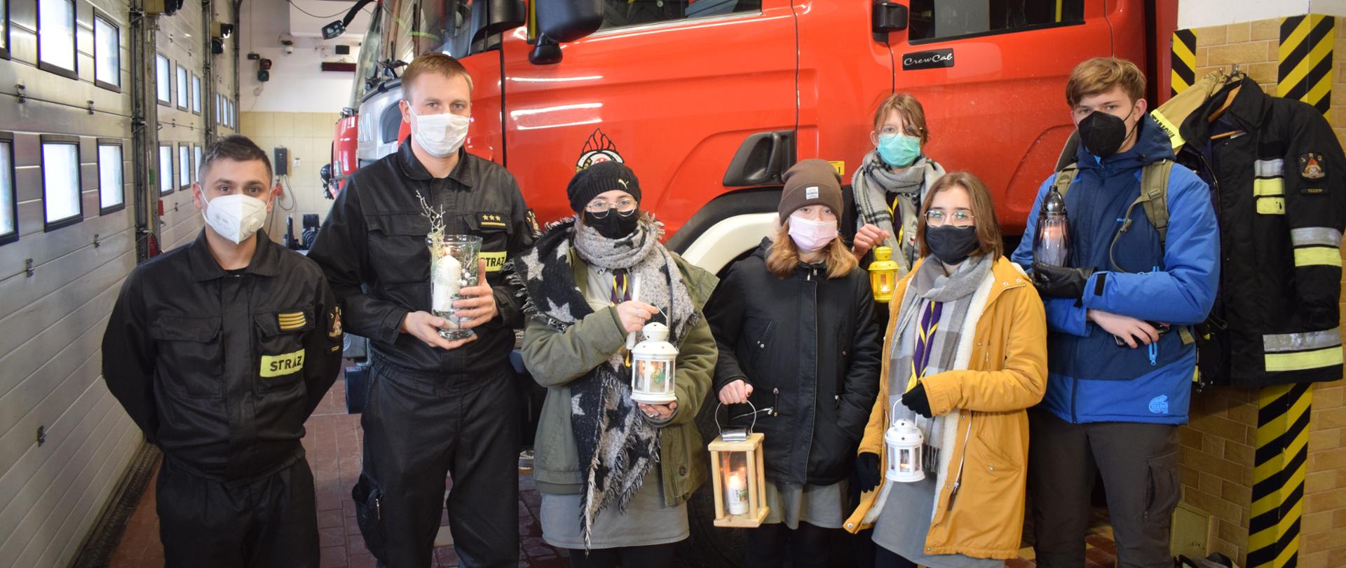 Garaż JRG Tczew. Na zdjęciu 7 osób. Od lewej dwóch strażaków. Następnie 5 harcerzy, trzymających lampiony z płonącym ogniem betlejemskim. W tle ciężki samochód gaśniczy.