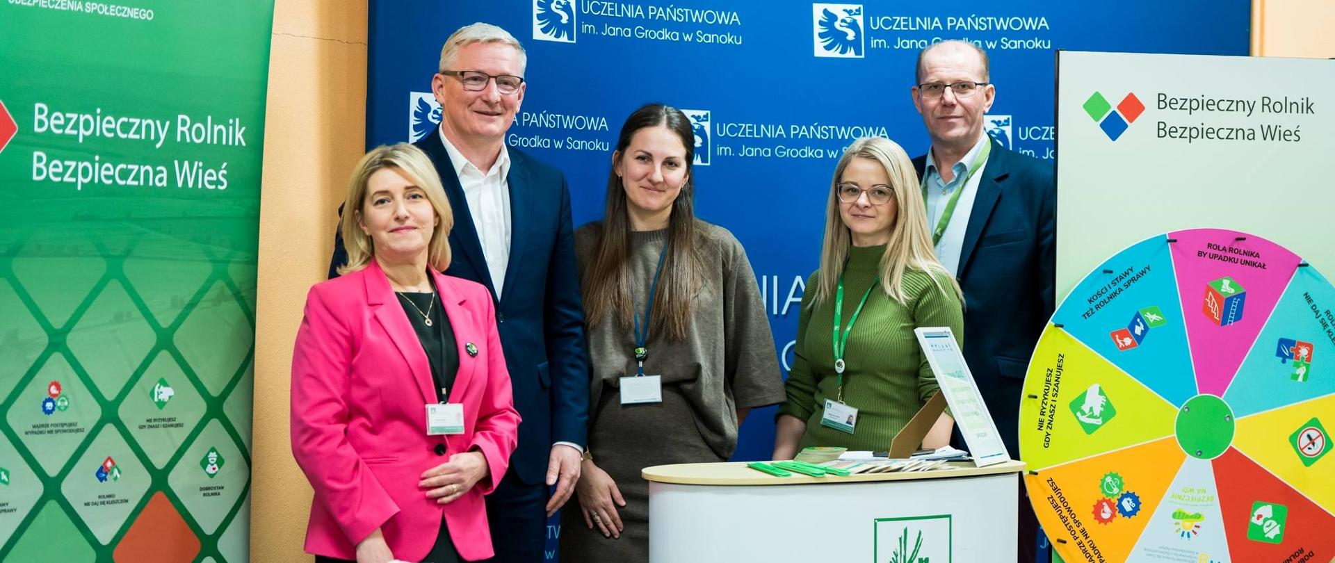 Trzy kobiety i dwóch mężczyzn stoją na tle roll-upów KRUS oraz Uczelni Państwowej im. Jana Grodka w Sanoku. Przed nimi znajduje się stół, na którym są wyłożone materiały prewencyjne Kasy. 