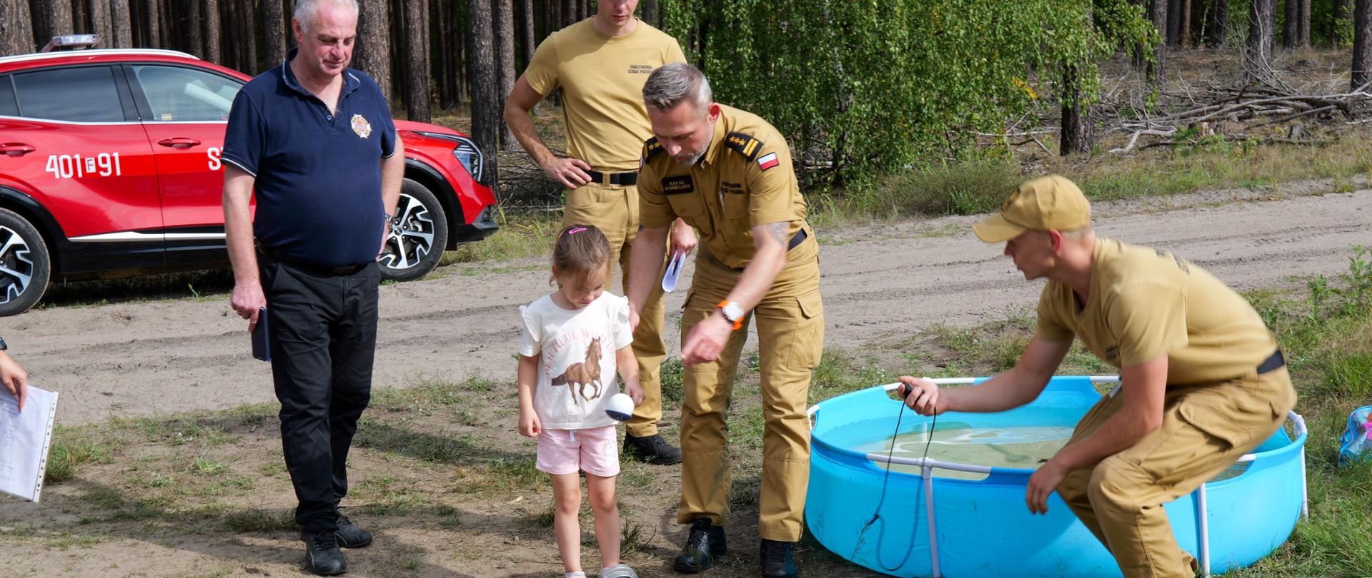 Strażacy w piaskowych mundurach PSP pomagają małej dziewczynce podczas konkurencji wodnej z wężem strażackim i basenem wypełnionym wodą; w tle widoczny czerwony samochód pożarniczy.