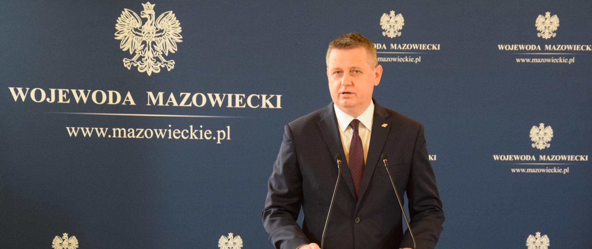 Mężczyzna ubrany w garnitur stoi za szarą mównicą i przemawia. W tle znajduje się granatowa ścianka z białymi orłami