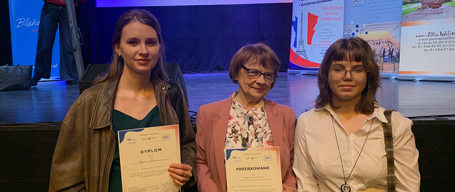 Trzy kobiety pozują na scenie, trzymając dyplomy i podziękowanie. W tle widoczny jest ekran z napisem „XXI edycji festiwalu” oraz banery informacyjne UMCS i KUL.
