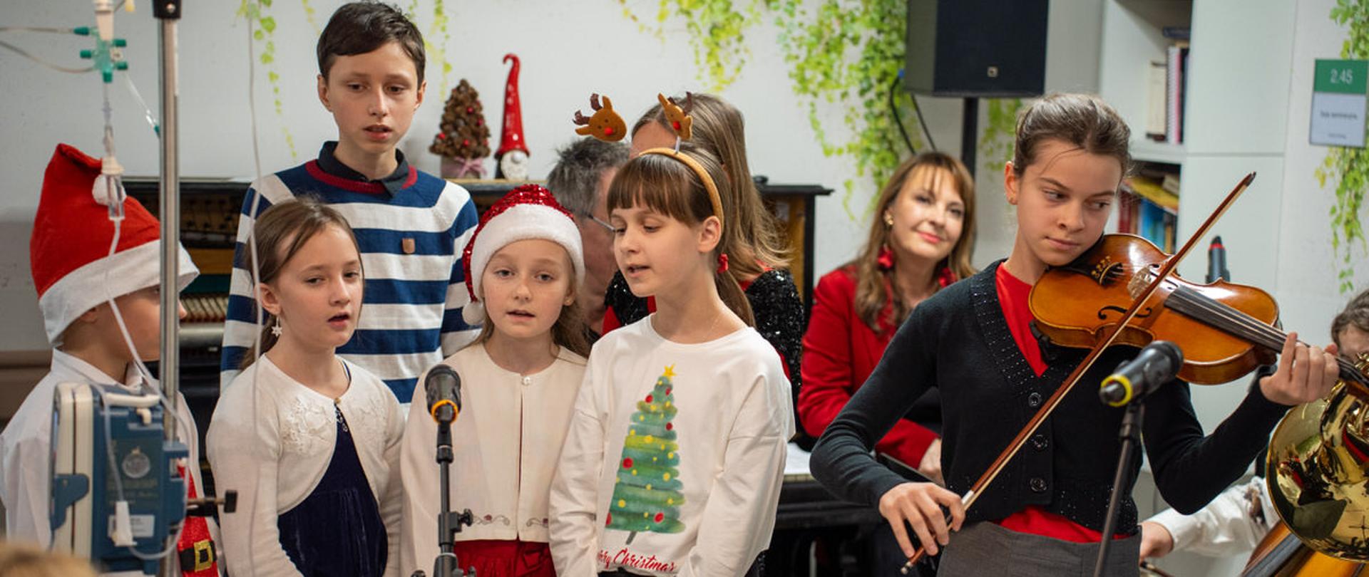 Na zdjęciu widoczni są uczniowie szkoły śpiewający i grający kolędy podczas koncertu w Klinice Onkologii Uniwersyteckiego Centrum Klinicznego Warszawskiego Uniwersytetu Medycznego. W tle siedzi towarzysząca im Pani Justyna Sieńczyłło.