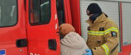 Strażak ochotniczej straży pożarnej w maseczce i rękawiczkach pomaga starszej osobie wsiąść do lekkiego pojazdu pożarniczego.