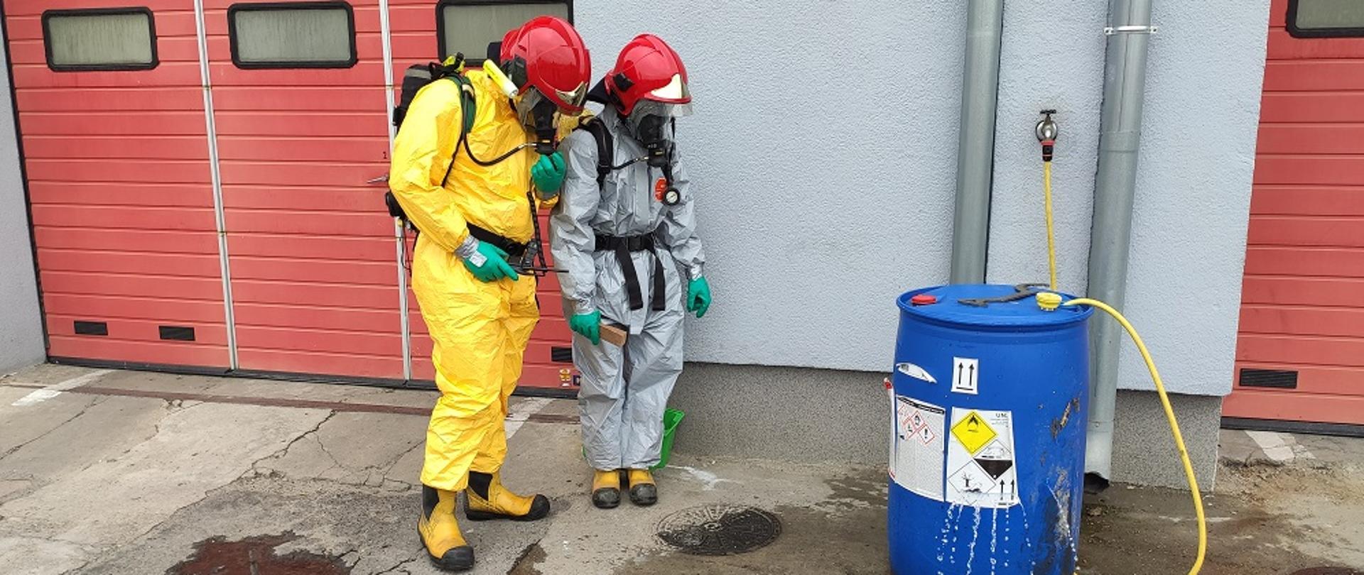 dwie osoby ubrane w ubraniach ochronne w hełmach stoi przed beczką z której wylatuje ciecz