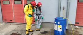 dwie osoby ubrane w ubraniach ochronne w hełmach stoi przed beczką z której wylatuje ciecz