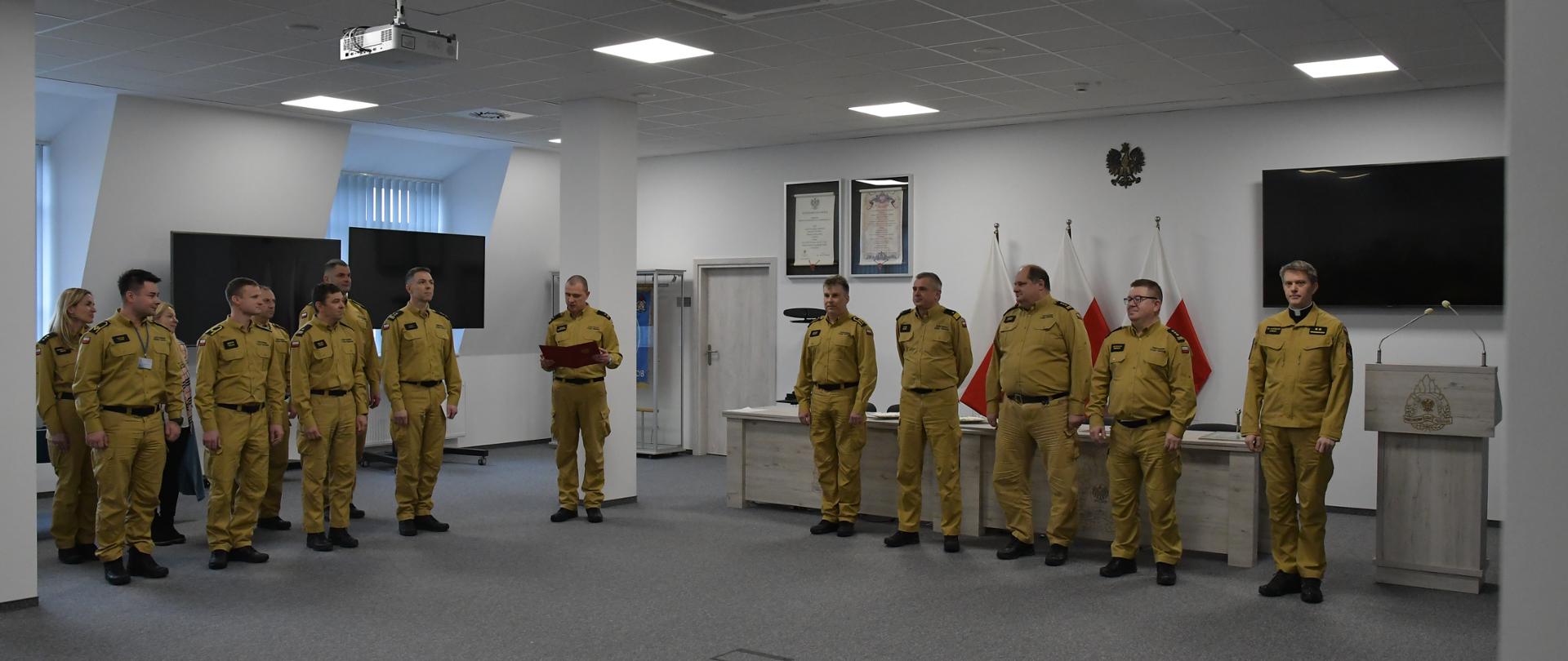 Kierownictwo Komendy Wojewódzkiej Państwowej Straży Pożarnej w Gdańsku wraz ze strażakiem oraz kapelanem stoją obok siebie za nimi ustawione są biurka i mównica trzy flagi Polski na ścianie wisi orzeł telewizor oraz rota ślubowania obok stoją strażacy jeden z nich trzyma teczkę i czyta.