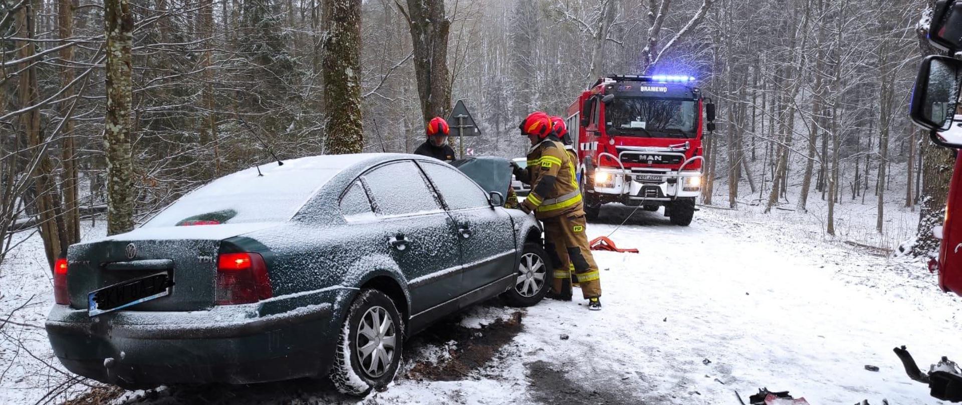 Zima, biało od śniegu. Z lewej strony stoi zielone auto osobowe przysypane śniegiem. Na drodze dużo śniegu. Dalej na drodze stoi samochód strażacki. Przy samochodzie osobowym strażacy w czerwonych hełmach.