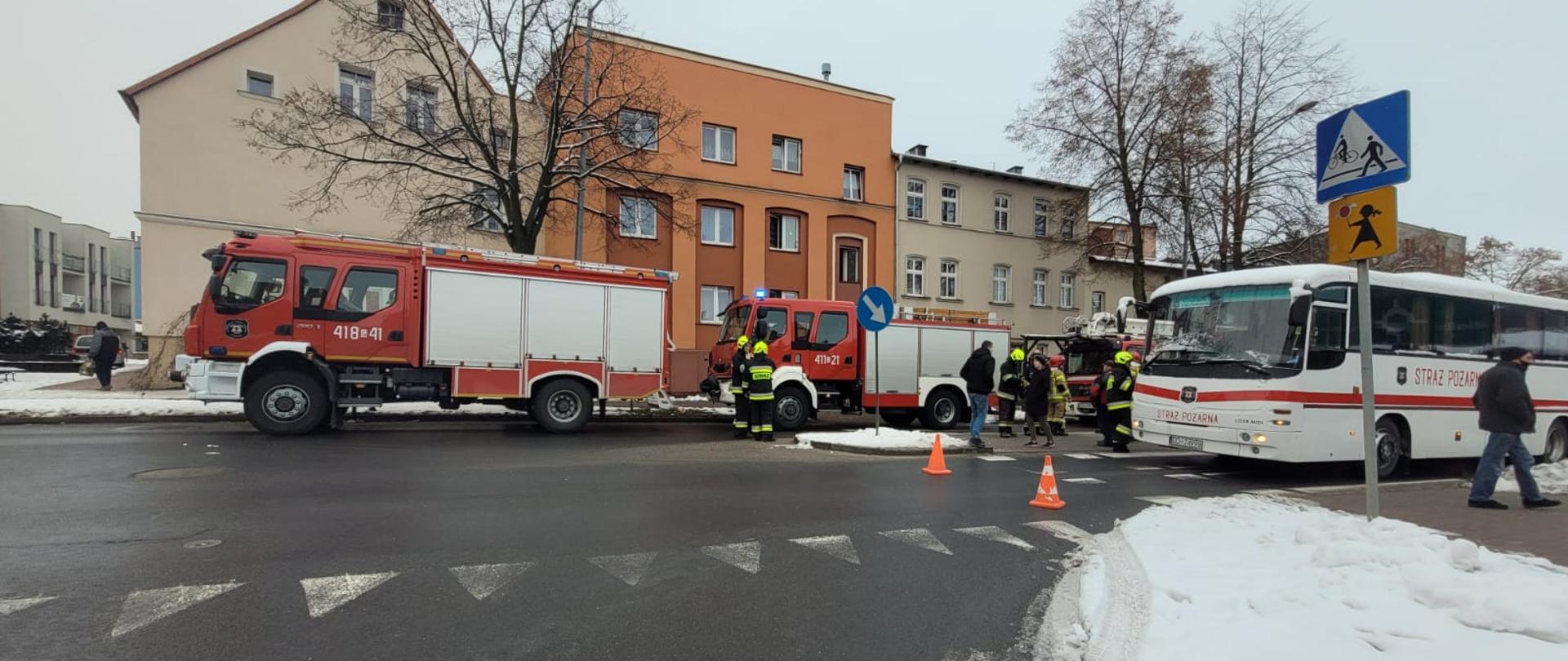 Pojazdy pożarnicze stojące na ulicy podczas ewakuacji mieszkańców z budynku. Widoczny autokar do którego ewakuują się mieszkańcy aby schronić się przed zimnem. 
