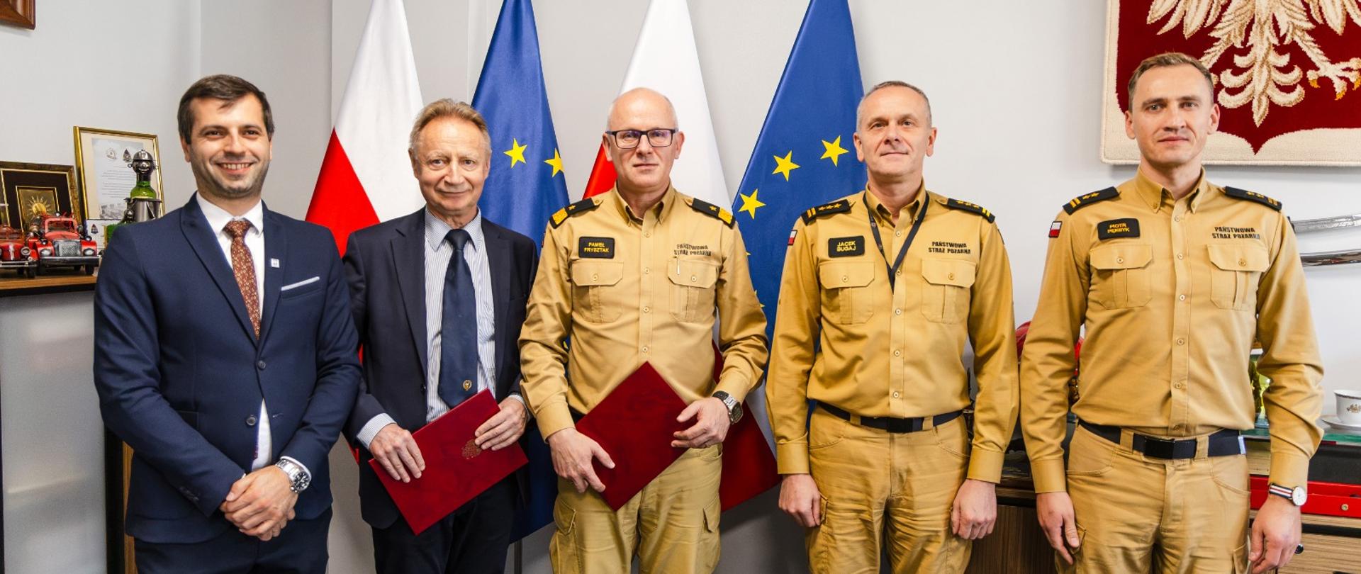Pięciu uczestników spotkania pozuje do wspólnego zdjęcia po podpisaniu dokumentów. Trzech strażaków PSP w umundurowaniu stoi obok dwóch mężczyzn w garniturach. W tle flagi Polski i Unii Europejskiej oraz godło Polski.