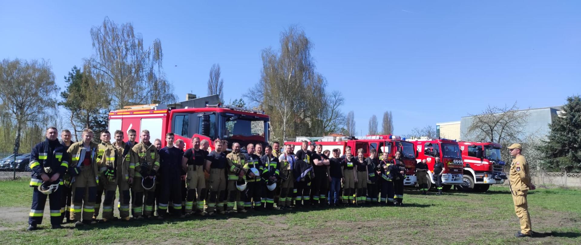 Zdjęcie w ciągu dnia. Niebo błękitne. Słonecznie. Na pierwszym planie ratownicy. Za ratownikami pojazdy ratowniczo-gaśnicze.