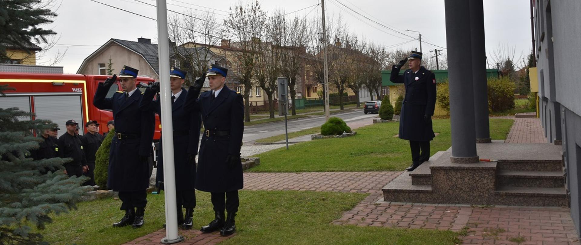 Na pierwszym planie widoczna jest grupa strażaków ubranych w ubrania koszarowe. Strażacy stoją w pozycji zasadniczej ustawieniu w dwuszeregu. Jeden z nich z lewej strony szeregu salutuje. Strażacy stoją na tle samochodu gaśniczego. Na drugim planie widoczny jest fragment ulicy oraz budynki mieszkalne.