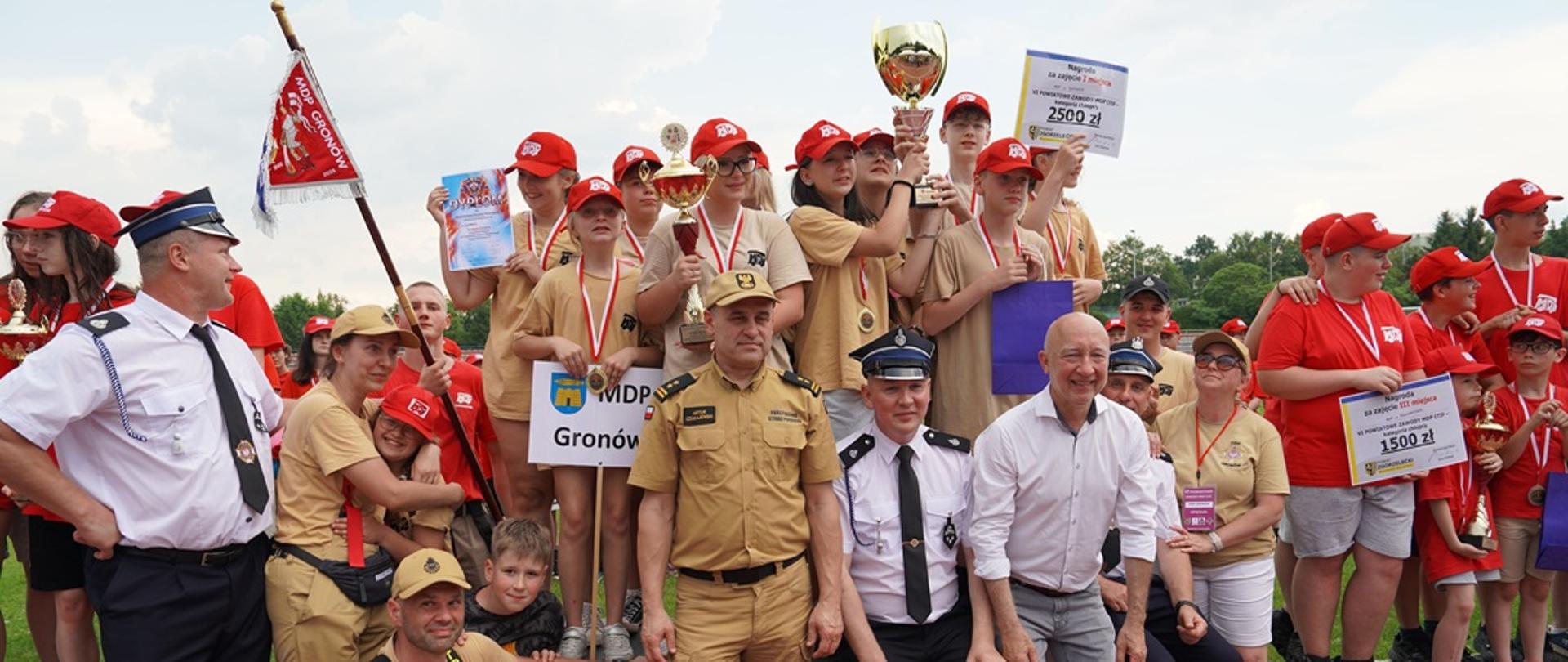 Grupka dzieci w koszulach w kolorze piaskowym stoi na podium. Dzieci na szyjach mają założone medale, a kilka z nich trzyma duży puchar. Przy podium stoi kilka osbó dorosłych, w tym funkcjonariusz PSP w piaskowym ubraniu służbowym i czapce z daszkiem, kilku druhów OSP ora jedna osoba cywilna.