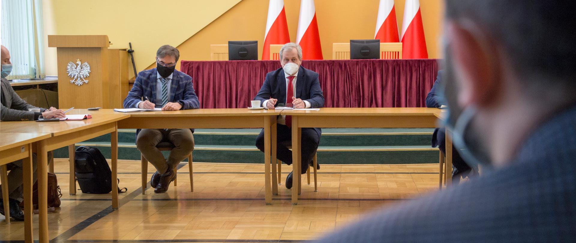 Spotkanie Zespołu Roboczego ds. zabezpieczenia Pilotażowego Punktu Szczepień Powszechnych w Opolu . Na pierwszym planie w głębi zdjęcia stół prezydialny, przy którym na środku siedzi wojewoda opolski. Wojewoda ma lekko uniesione nad blatem stołu ręce, na twarzy ma maseczkę medyczna, spogląda na uczestników spotkania. Obok niego - po lewej stronie kadru, siedzi dyrektor opolskiego NFZ. Mężczyzna pochylony jest nad karta papieru. Na nosie ma okulary optyczne. Po prawej stronie kadru widać rozmytą postać - a konkretniej osobę, siedzącą tyłem do obiektywu. W tle flagi biało-czerwone. 