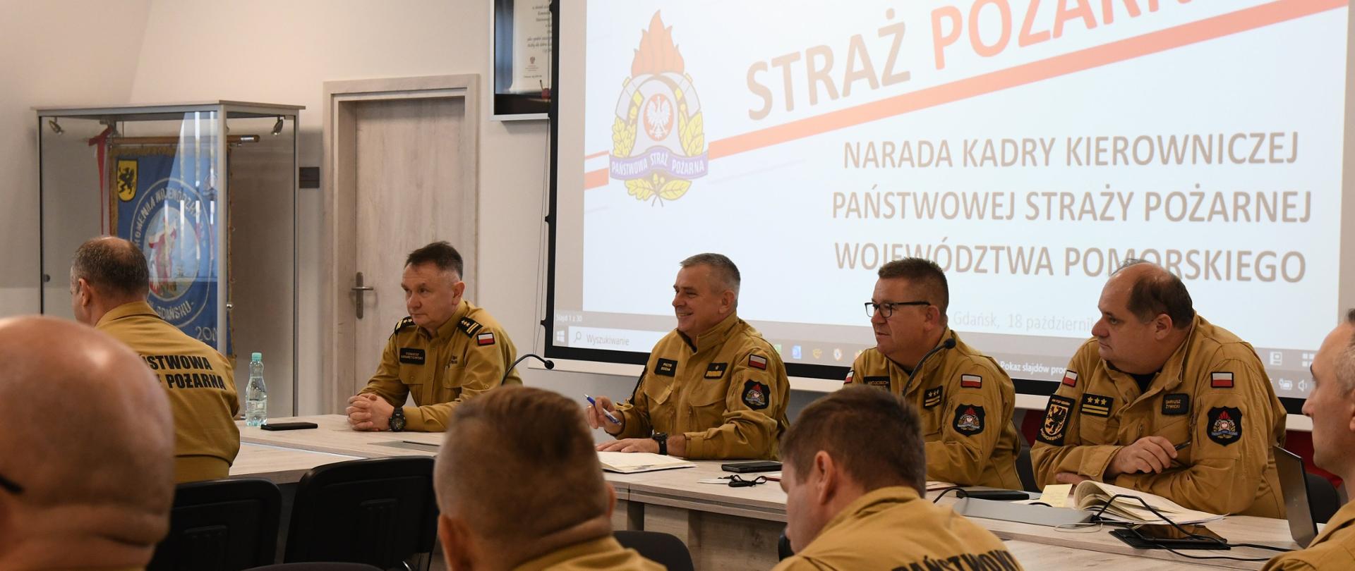 Funkcjonariusze Państwowej Straży Pożarnej w mundurach koloru musztardowego siedzą za biurkami obok siebie za nimi na ekranie wyświetlany jest napis narada kadry kierowniczej Państwowej Straży Pożarnej województwa pomorskiego obok w witrynie umieszczony jest sztandar Komendy Wojewódzkiej Państwowej Straży Pożarnej w Gdańsku. 
