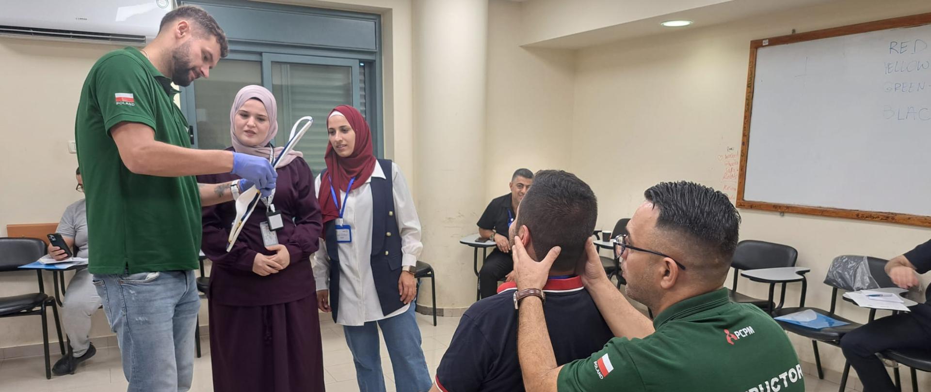 Mężczyzna w zielonym tshircie z napisem Poland pokazuje coś na sprzęcie medycznym , dwie kobiety w chustach się temu przyglądają. Drugi mężczyzna w zielonym tshircie z napisem instructor obejmuje głowę mężczyzny siedzącego przed nim