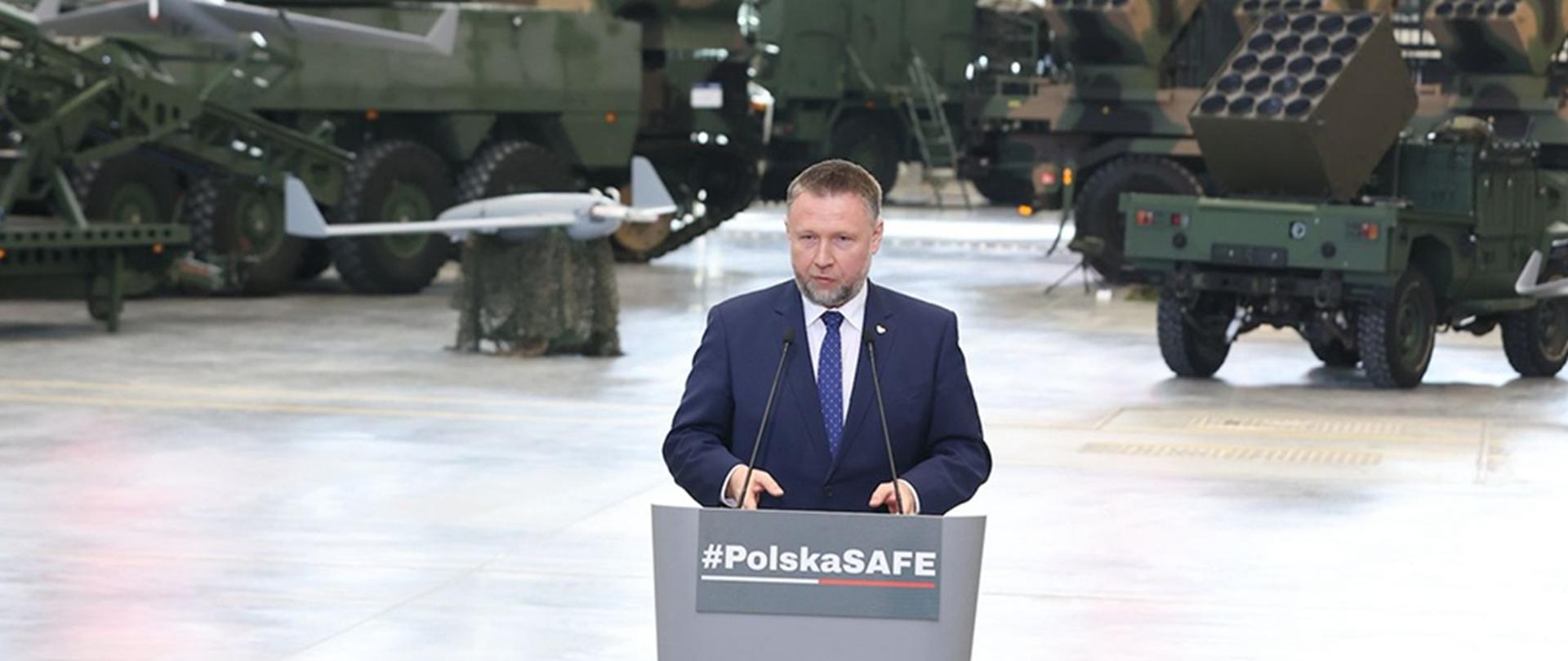 Minister Marcin Kierwiński na mównicy podczas konferencji prasowej w hali z pojazdami wojskowymi.