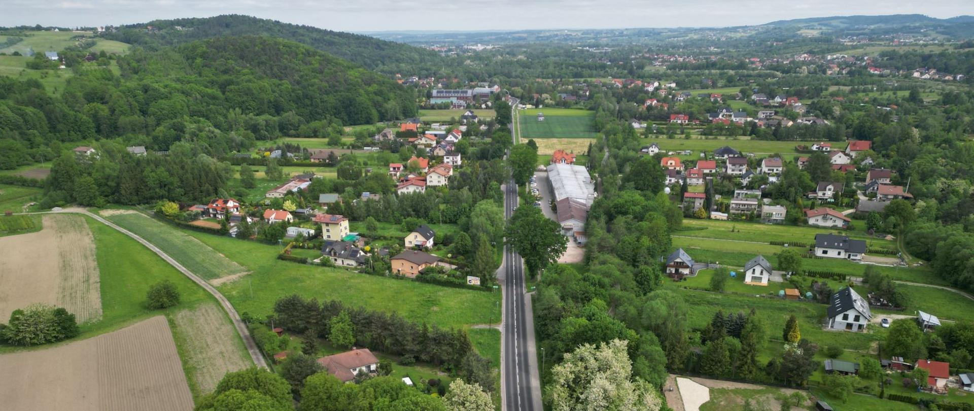 Widok z drona na drogę krajową 28 (odc. Wadowice-Sucha Beskidzka), widoczna zabudowa jednorodzinna zatopiona w zieleni oraz tereny leśne. W tle widoczne Beskidy.