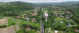 Widok z drona na drogę krajową 28 (odc. Wadowice-Sucha Beskidzka), widoczna zabudowa jednorodzinna zatopiona w zieleni oraz tereny leśne. W tle widoczne Beskidy