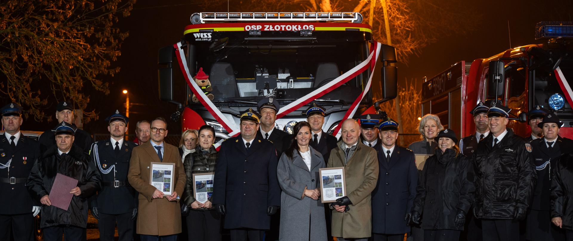 Zdjęcie grupowe uczestników uroczystości, stojących przed czerwonym samochodem strażackim z napisem "OSP ZŁOTOKŁOS". W tle ciemne nocne niebo oraz drzewa oświetlone uliczną lampą. 