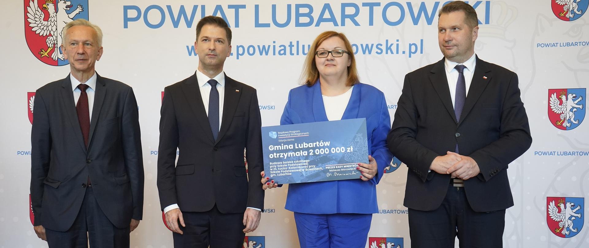 Pod ścianką z napisem Powiat Lubartowski stoi minister Czarnek, dwóch mężczyzn w garniturach i kobieta w niebieskim ubraniu która trzyma tabliczkę z napisem Gmina Lubartów.