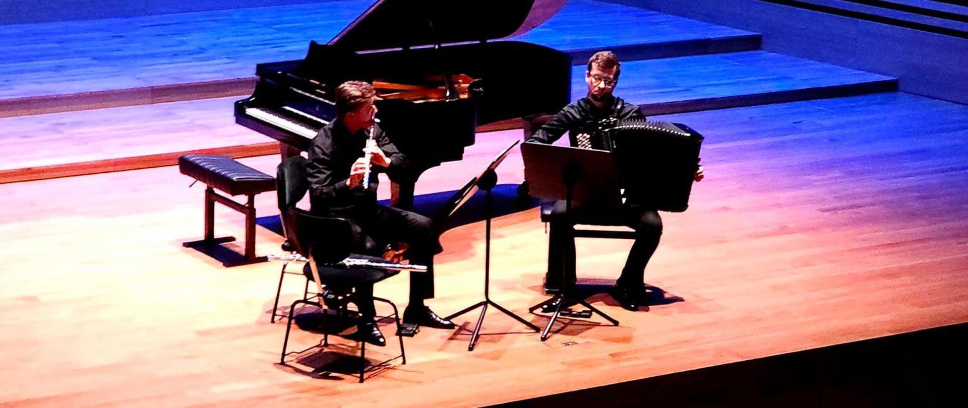 Dwaj mężczyźni siedząc na krzesłach grają na flecie i akordeonie, za nimi stoi fortepian.