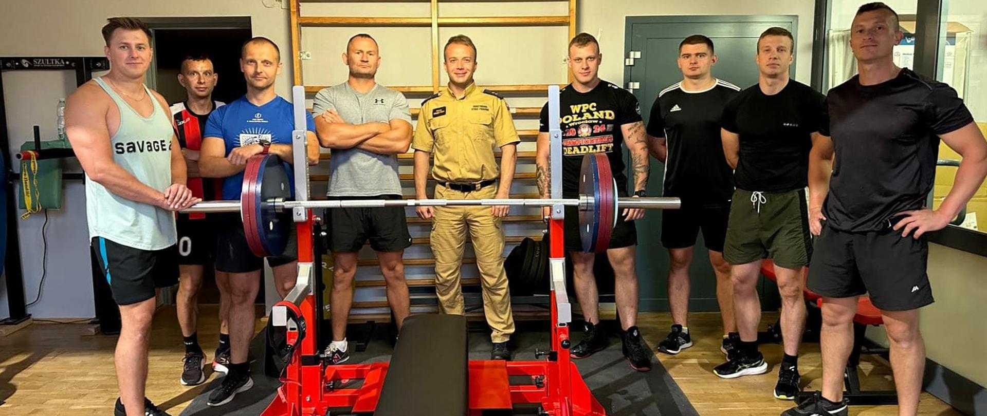 Zdjęcie grupowe uczestników szkolenia na tle ławki ze sztangą