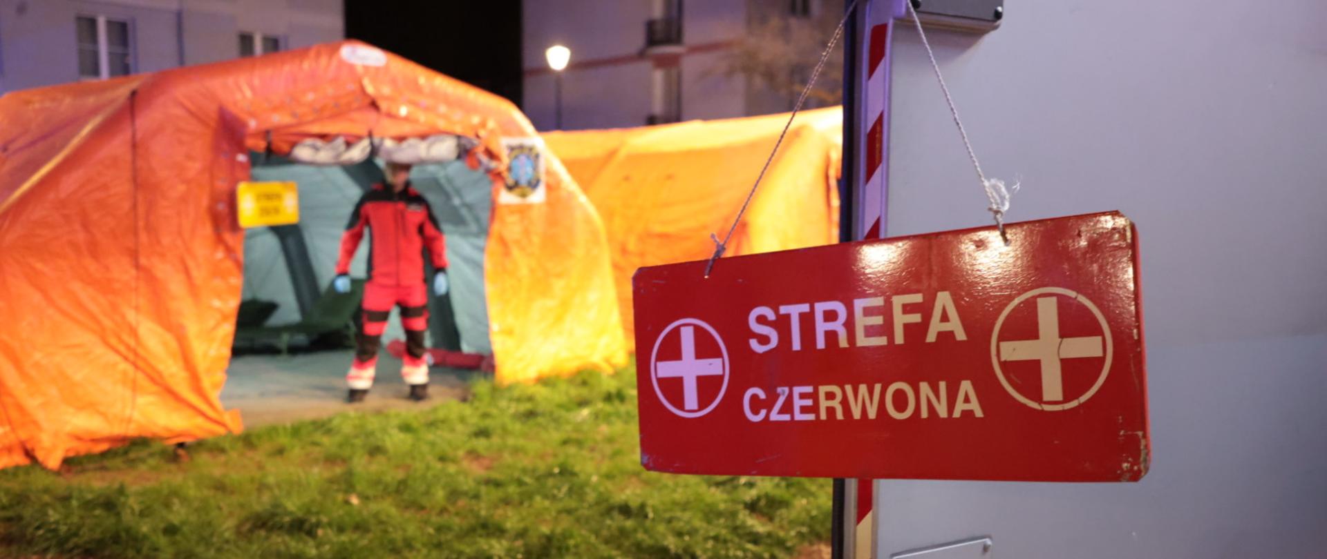 Na klamce do drzwi samochodu wisi tabliczka czerwona z napisem STREFA CZERWONA, w tle dwa namioty do dekontaminacji, w wejsciu do jednego z nich widać stojącego strażaka