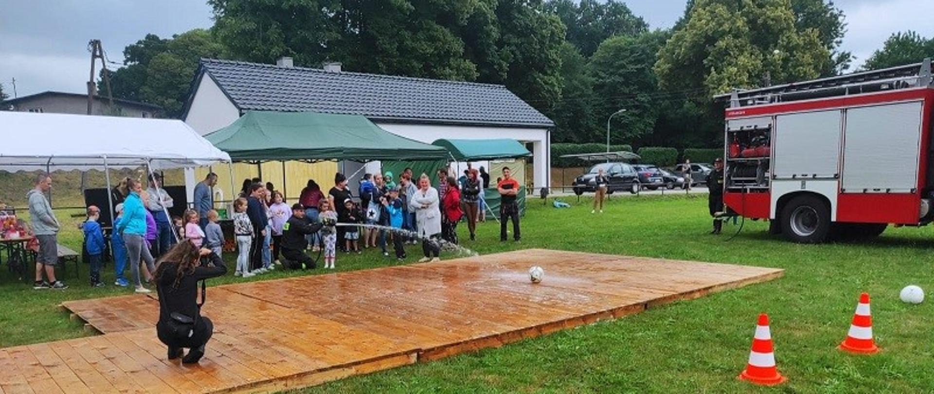 „Bezpieczne Wakacje” w miejscowości Bukówko. Zdjęcie przedstawia pojazd strażacki oraz dzieci grające w piłkę wodną. Dziewczynka za pomocą prądownicy wodnej podaje strumień wody na piłkę próbując skierować ją do przestrzeni pomiędzy dwoma pachołkami drogowymi. W głębi zdjęcia pod namiotami uczestnicy festynu