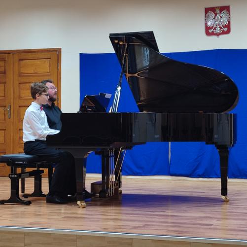 Nauczyciel ubrany na czarno i uczeń w białej koszuli, czerwonej muszce oraz czarnych spodniach graja na fortepianie na cztery ręce. W tle niebieska kotara. 
