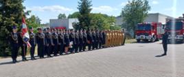 Pododdział Strażków podczas uroczystości zgromadzeni na placu. w tle samochody pożarnicze