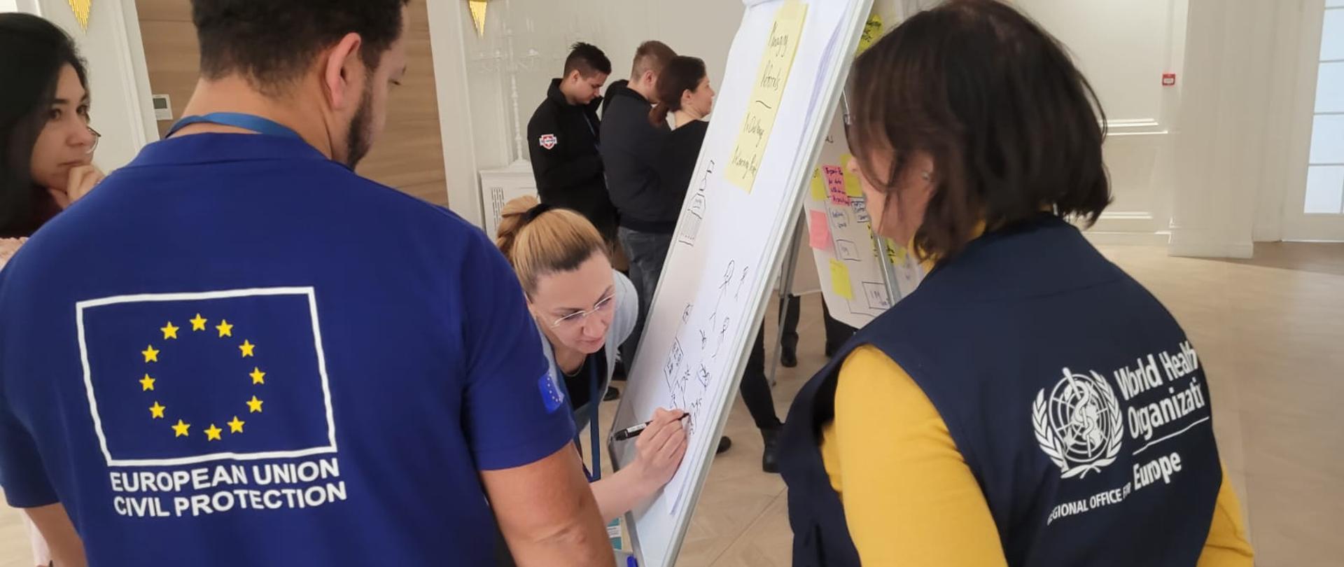 Widok z tyłu na postać dwóch osób, po prawo na ubraniu kobiety napis World Health Organization, po lewej na koszulce mężczyzny napis EUROPEAN UNION CIVIL PROTECTION