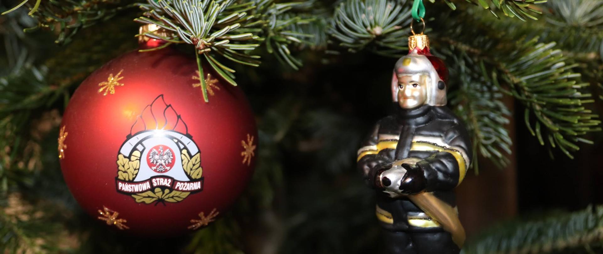 Po lewej bąbka choinkowa w kolorze czerwonym z logo PSP. Po prawej bąbka w kształcie strażaka w ubraniu bojowym z wężem. W tle choinka