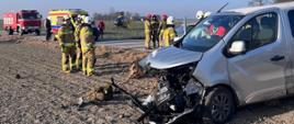 Na zdjęciu uszkodzony samochód typu bus do przewozu osób niepełnosprawnych. Koło pojazdu dwóch strażaków w umundurowaniu specjalnym, którzy usuwają skutki zdarzenia. W tle samochód strażacki, karetka pogotowia oraz śmigłowiec lotniczego pogotowia ratunkowego.